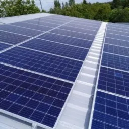 Błękitne panele fotowoltaiczne na jasnym, metalowym dachu, widok z góry, z zielenią drzew w tle.