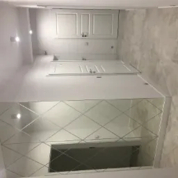 Nowoczesny korytarz z dużym lustrem z dekoracyjnymi szlifami w romby, białymi drzwiami i płytkami podłogowymi imitującymi kamień.