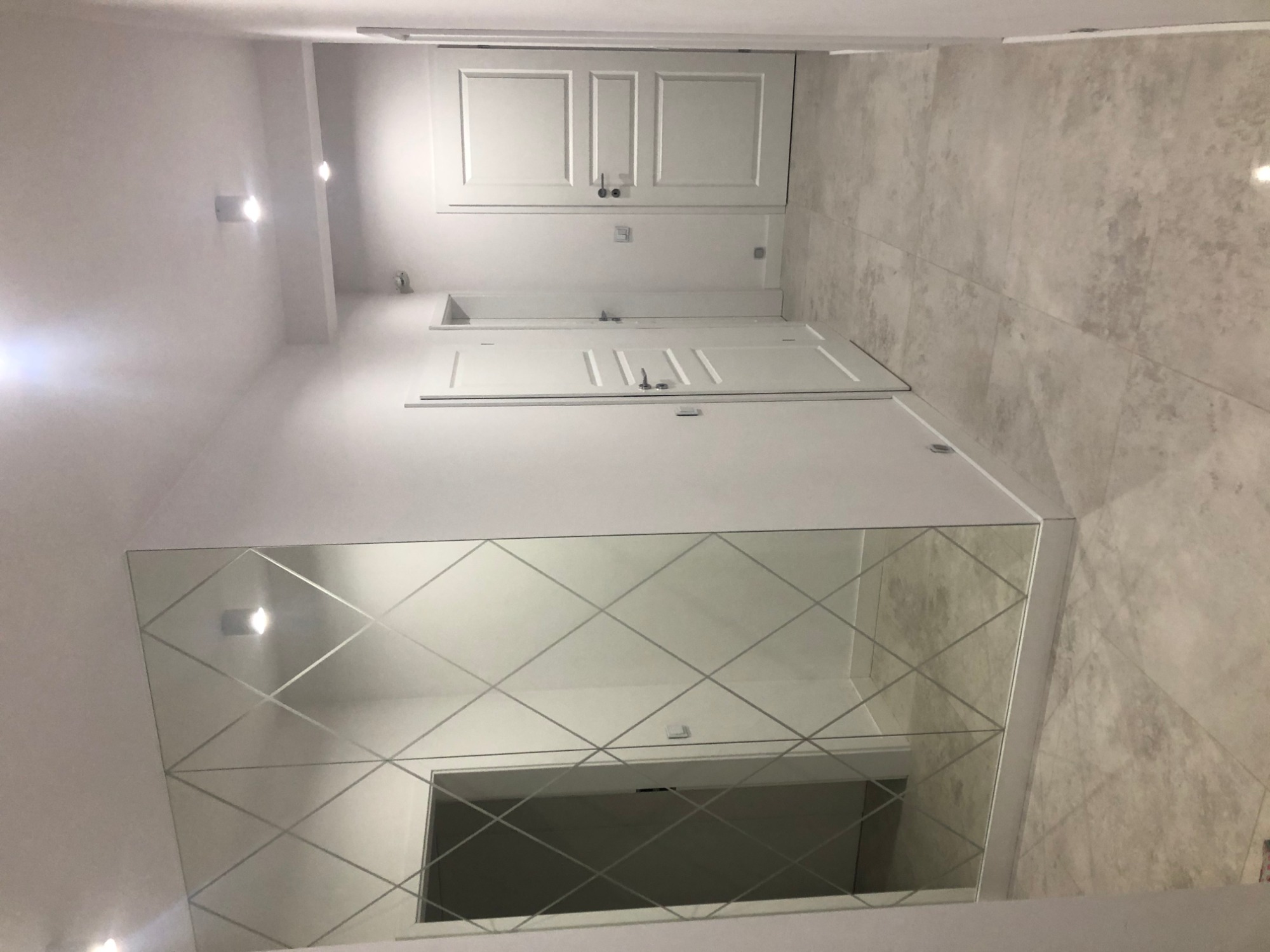 Nowoczesny korytarz z dużym lustrem z dekoracyjnymi szlifami w romby, białymi drzwiami i płytkami podłogowymi imitującymi kamień.