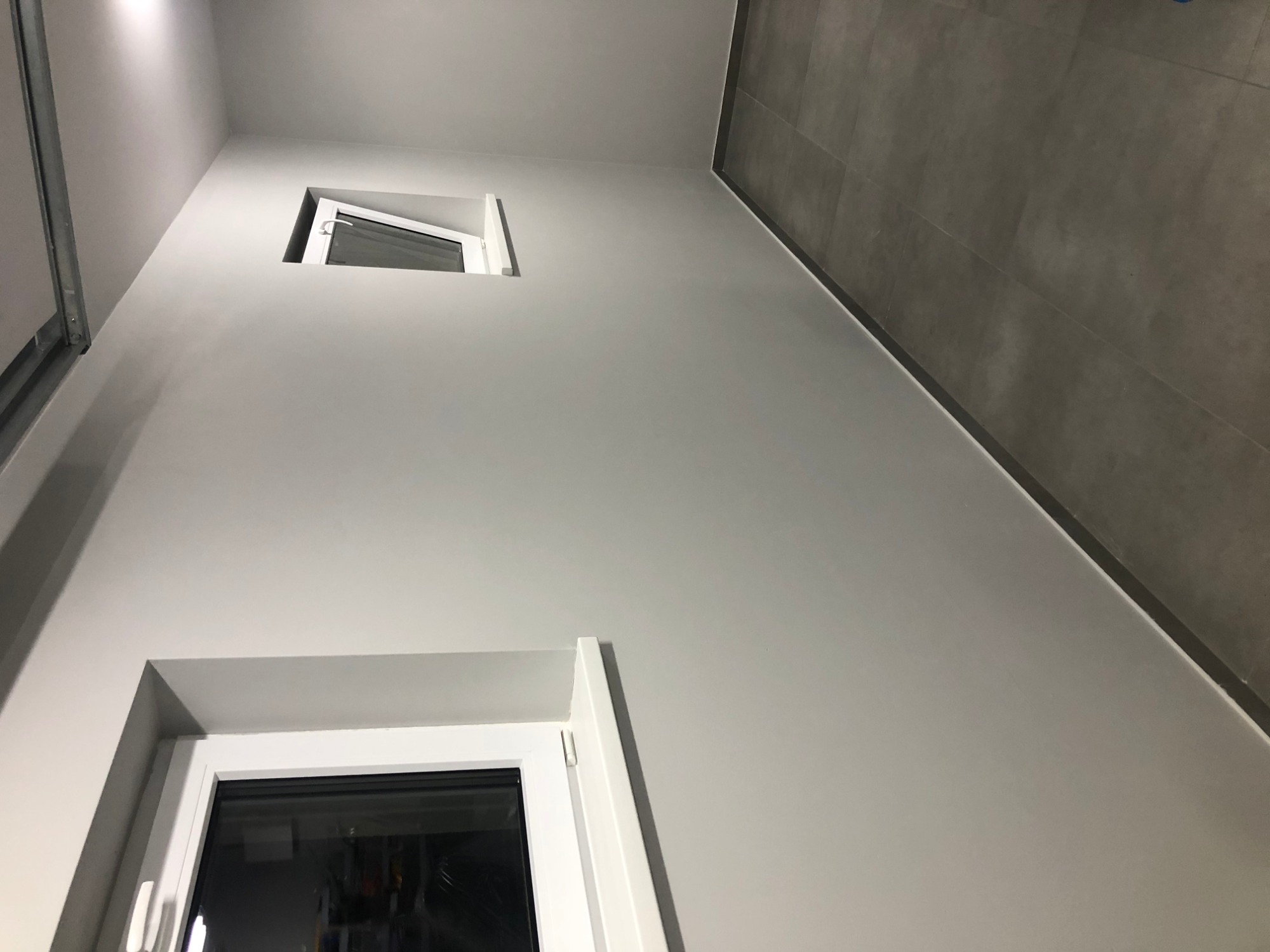 Świeżo pomalowane na szaro wnętrze z dwoma oknami, widoczne płytki podłogowe w odcieniach szarości.