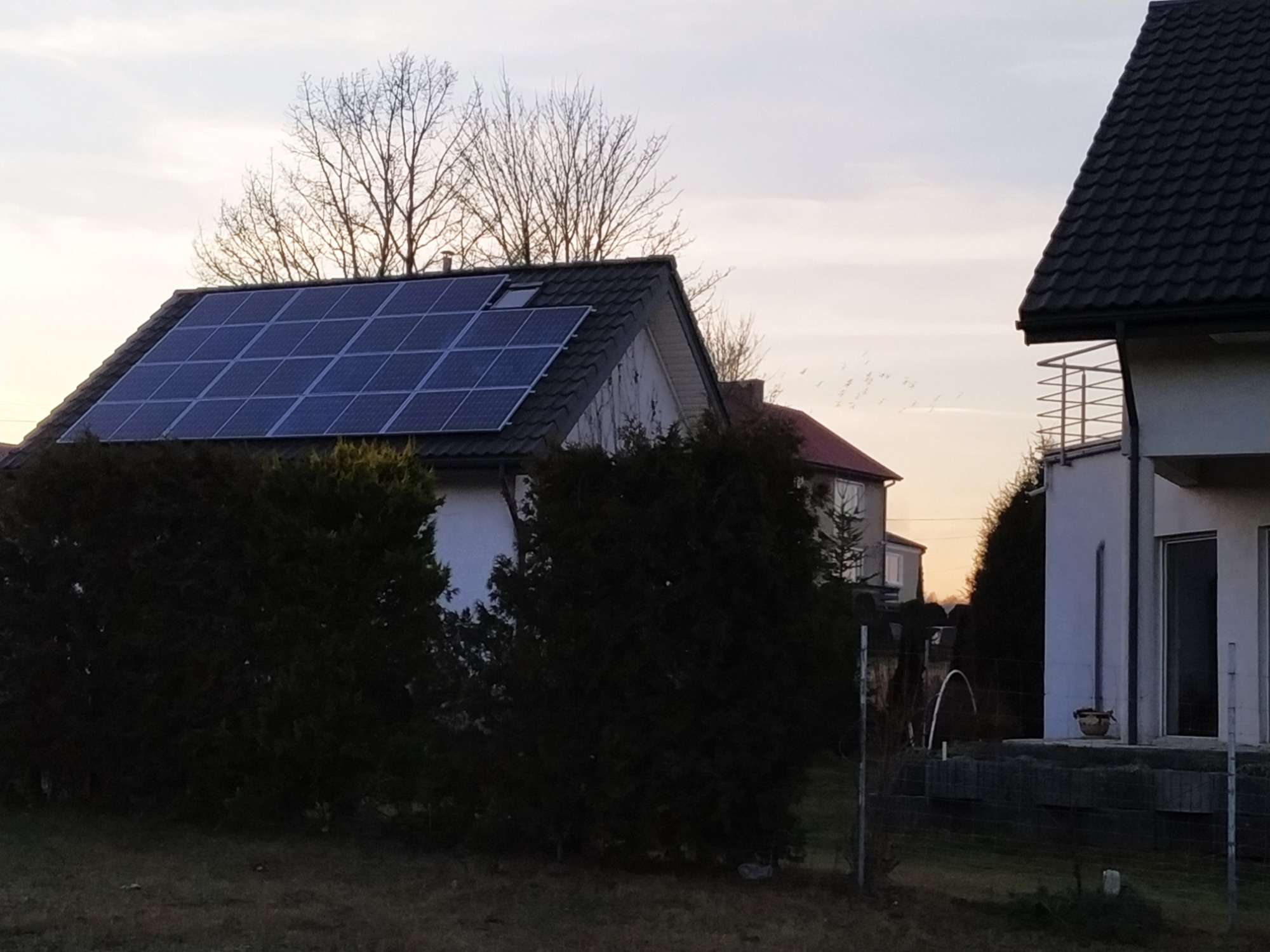 Dom jednorodzinny z panelami słonecznymi na dachu, widoczne z poziomu gruntu, w tle zachodzące słońce i przelatujące ptaki.