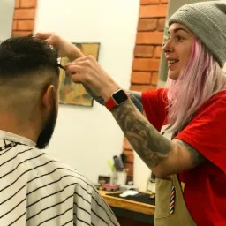 Kobieta z różowymi włosami, tatuażami na rękach i w szarej czapce strzyże brodatego mężczyznę w pelerynie w paski, używając grzebienia i nożyczek w salonie z ceglaną ścianą.