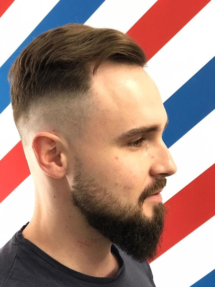 Mężczyzna z modnym cieniowaniem włosów i zadbaną brodą na tle ściany w czerwono-biało-niebieskie pasy.