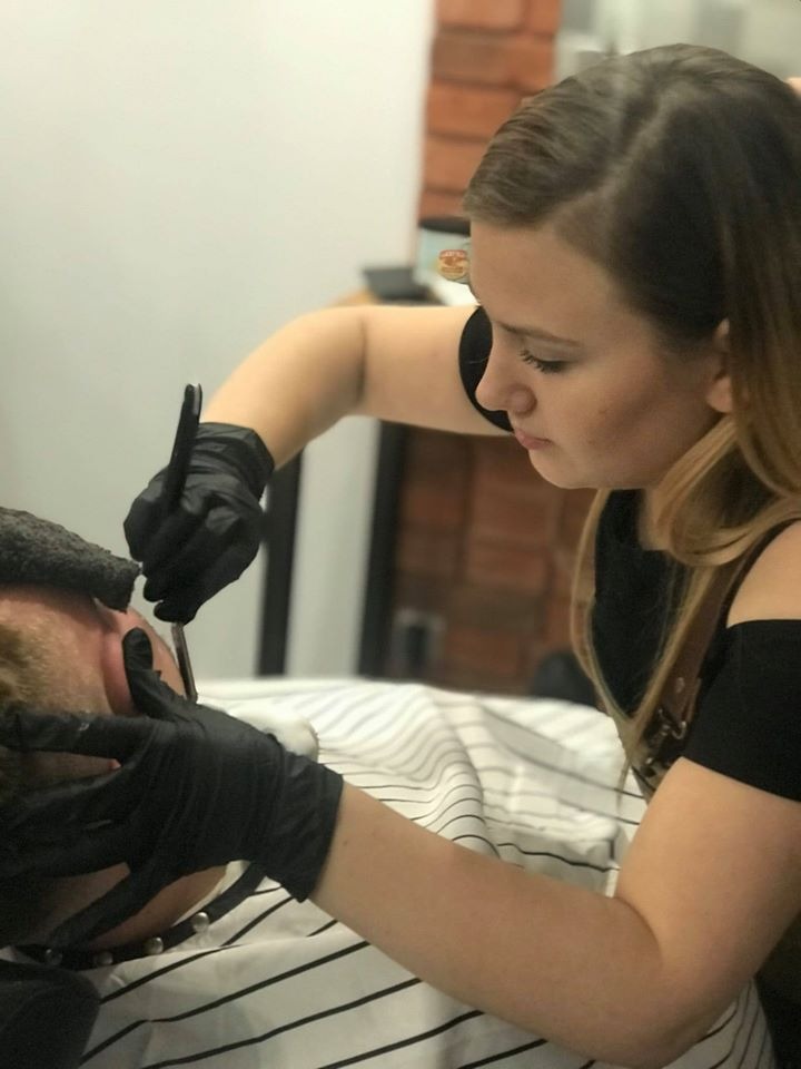 Kobieta w czarnych rękawiczkach goli twarz mężczyzny brzytwą w salonie, widoczny pasek na szyi klienta i fragment ceglanej ściany w tle.