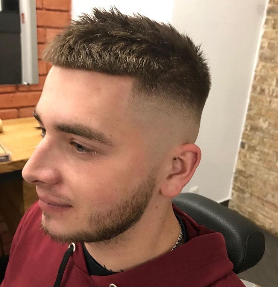 Młody mężczyzna z krótko ostrzyżonymi, brązowymi włosami, świeżo po wizycie u fryzjera, widoczny z profilu w fotelu fryzjerskim, z delikatnym zarostem i srebrnym łańcuszkiem na szyi.