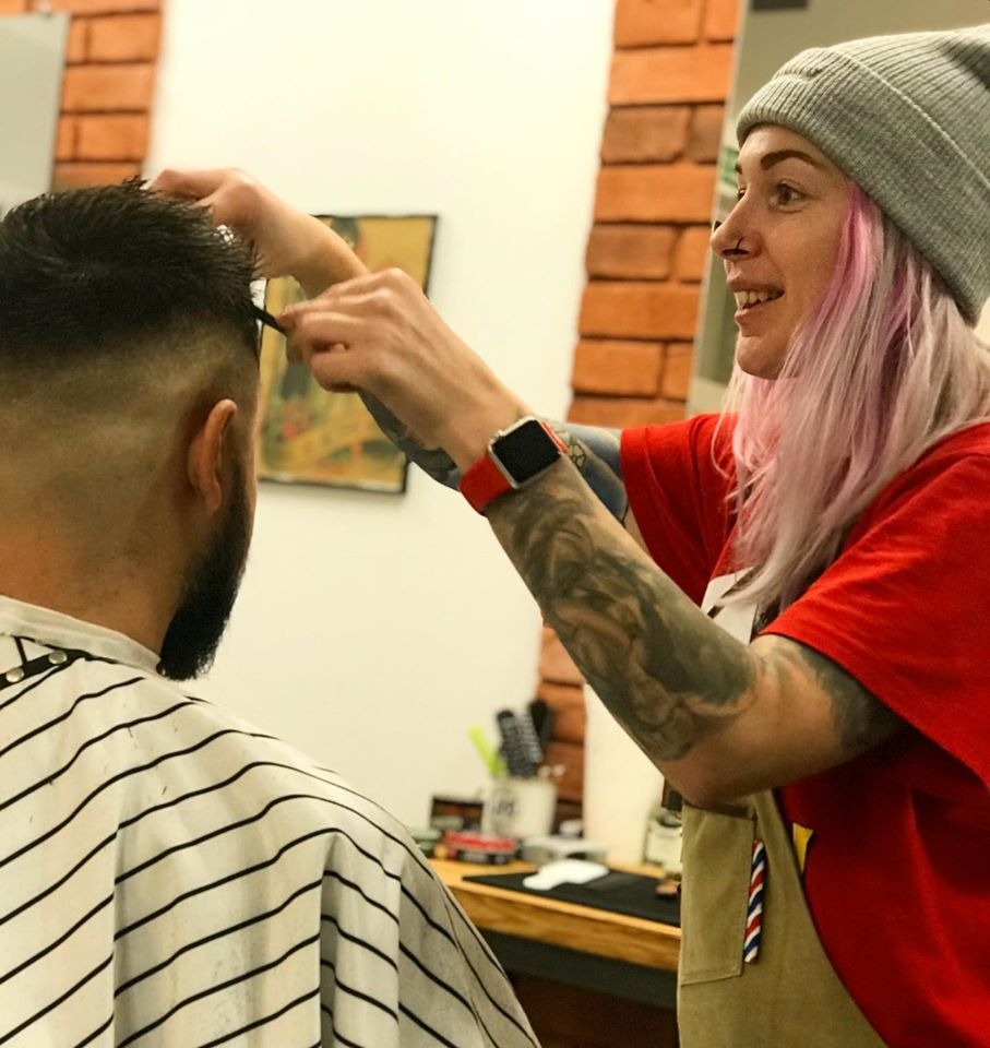 Kobieta z różowymi włosami, tatuażami na rękach i w szarej czapce strzyże brodatego mężczyznę w pelerynie w paski, używając grzebienia i nożyczek w salonie z ceglaną ścianą.
