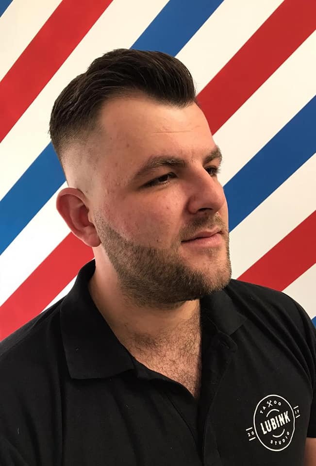 Mężczyzna z modną fryzurą typu fade i starannie przystrzyżoną brodą, na tle ściany w czerwono-biało-niebieskie pasy, w czarnej koszuli polo z logo Lubink Studio.