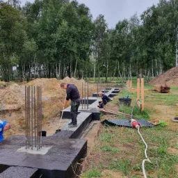 Prace izolacyjne fundamentów: robotnicy zabezpieczają betonowe ławy fundamentowe czarną izolacją bitumiczną, widoczne wystające pręty zbrojeniowe, wykop, oraz zaplecze budowy w tle.