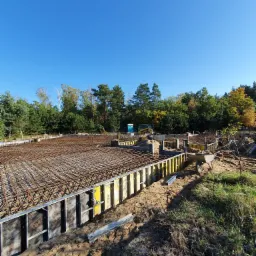 Przygotowanie terenu pod budowę: widok na szalunki fundamentowe wypełnione siatką zbrojeniową, w tle drzewa i błękitne niebo.
