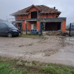 Surowy stan budowy domu jednorodzinnego z częściowo ukończoną konstrukcją dachu, widoczny samochód dostawczy i błotniste podłoże.