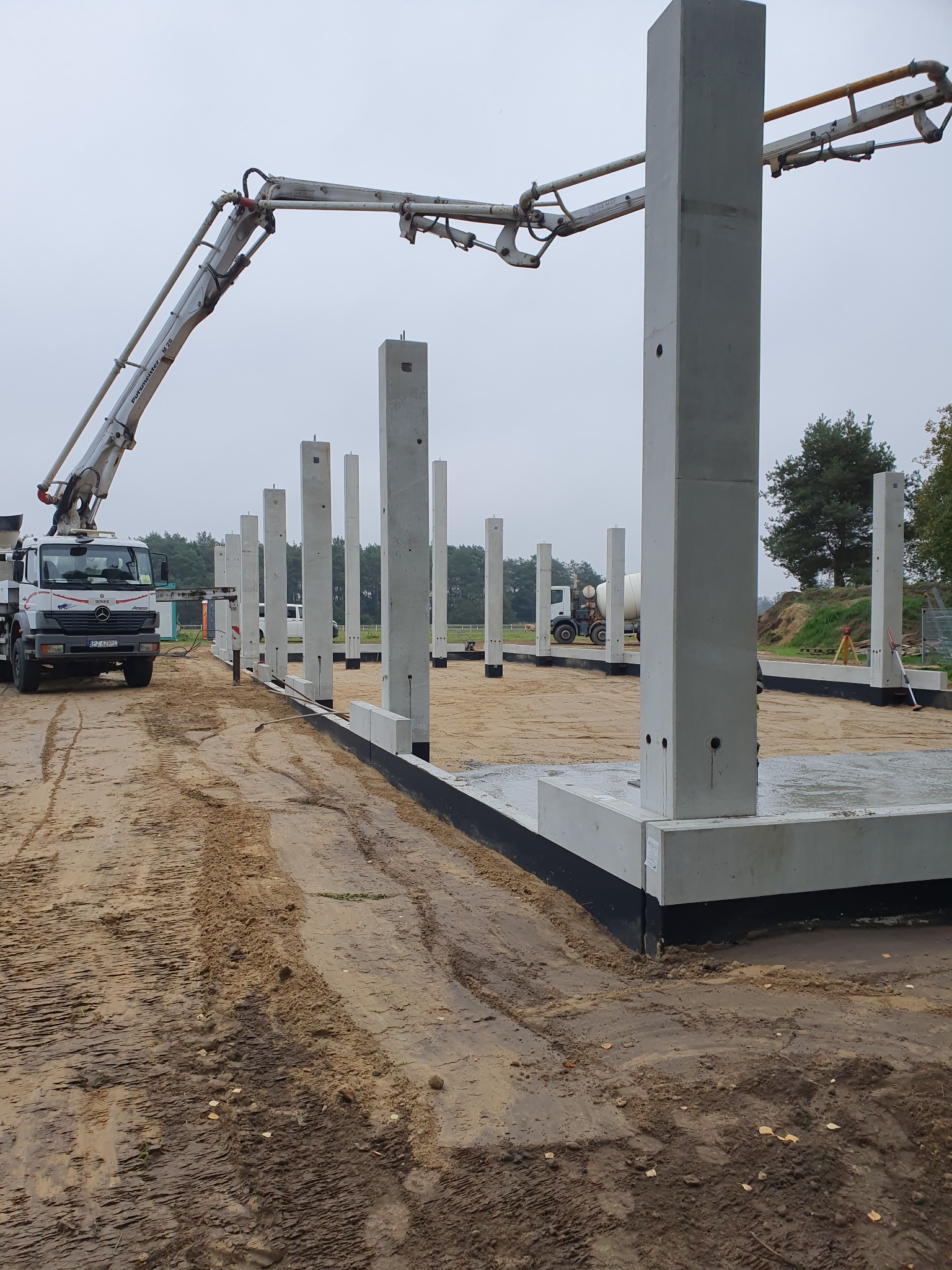Wznoszenie konstrukcji budynku: widok na betonowe słupy fundamentowe na przygotowanym terenie, z pompą do betonu i betoniarką w tle, w pochmurny dzień.