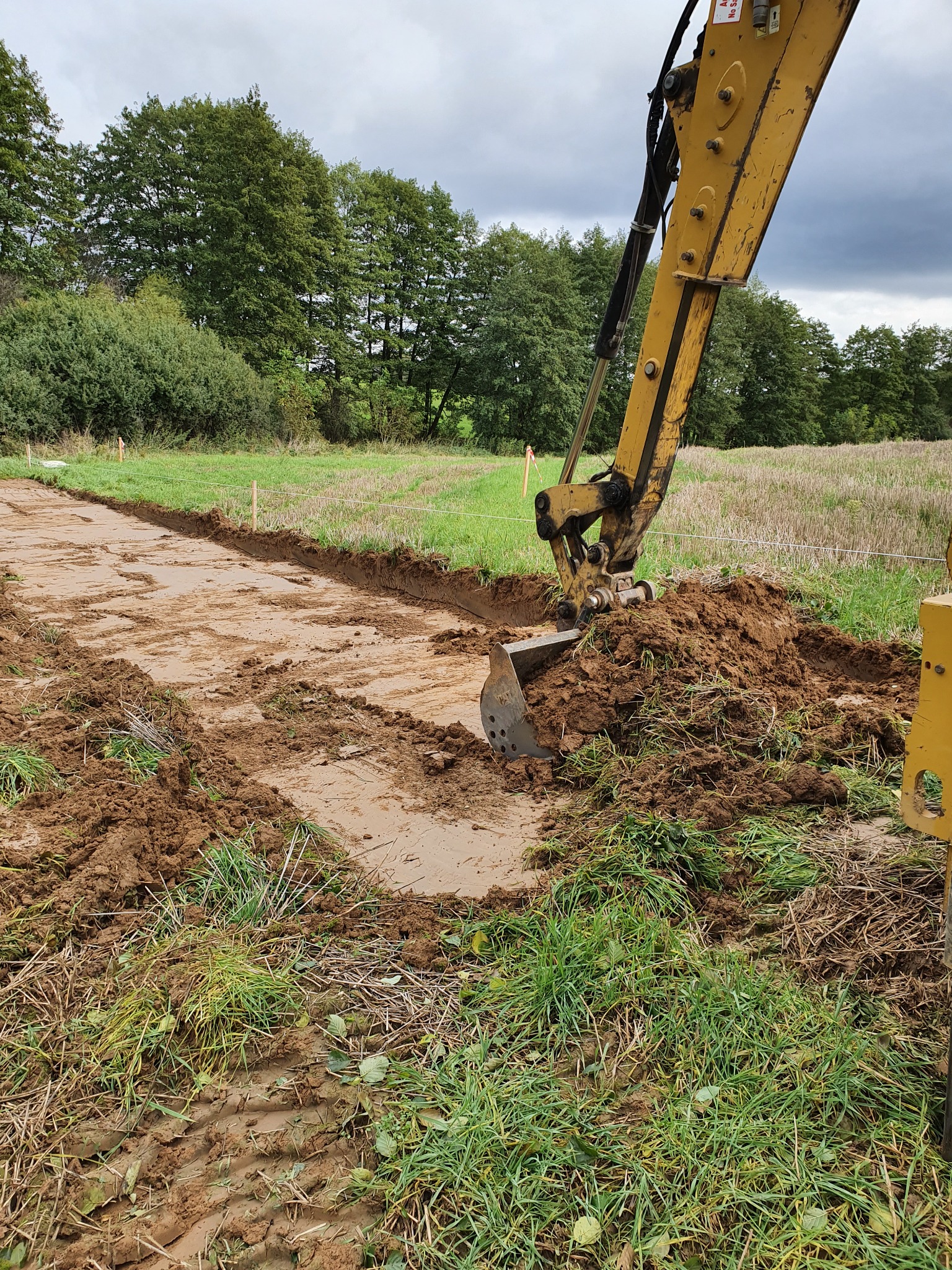 Żółta koparka w trakcie wykonywania wykopu w ziemi, widoczne świeżo wykopane koryto, trawa i drzewa w tle.