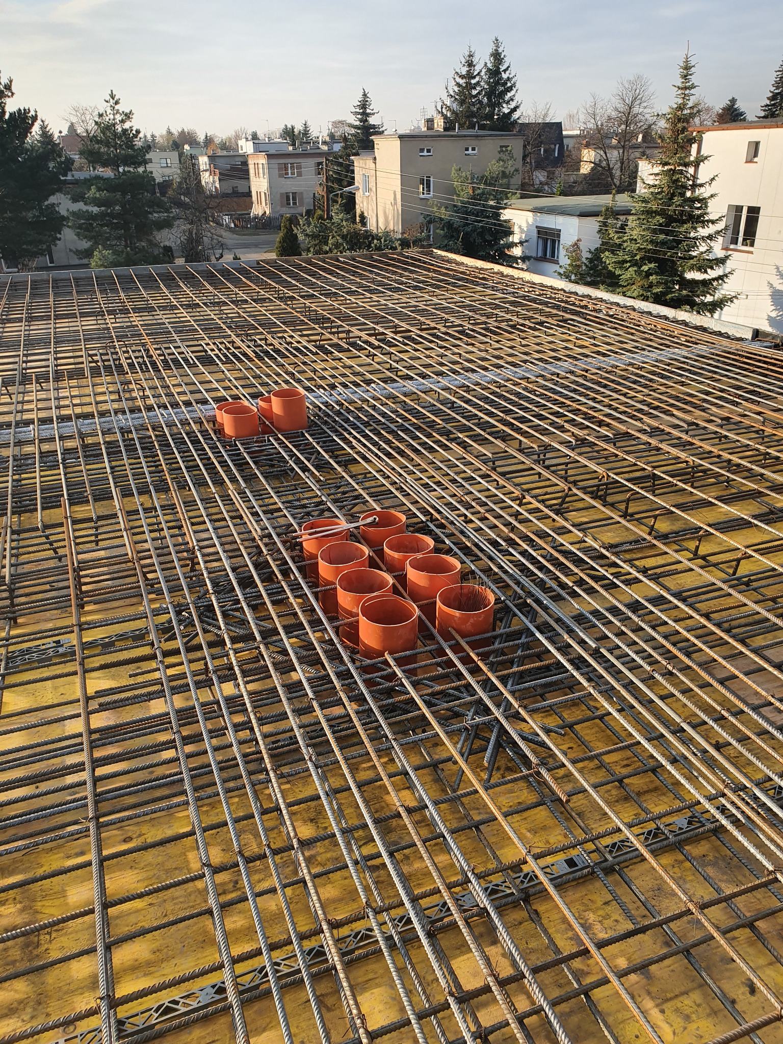 Zbrojenie stropu z widocznymi pomarańczowymi rurami osłonowymi na tle zabudowy miejskiej.