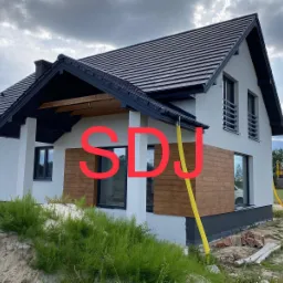 Nowoczesny dom jednorodzinny z białą elewacją, fragmentem drewnianej okładziny, czarnym dachem i orynnowaniem, widoczne elementy rusztowania i teren budowy.