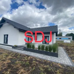 Nowoczesny dom jednorodzinny z białymi ścianami i ciemną dach&oacute;wką, z utwardzonym terenem wok&oacute;ł, schodami i rabatami z ozdobnymi trawami.