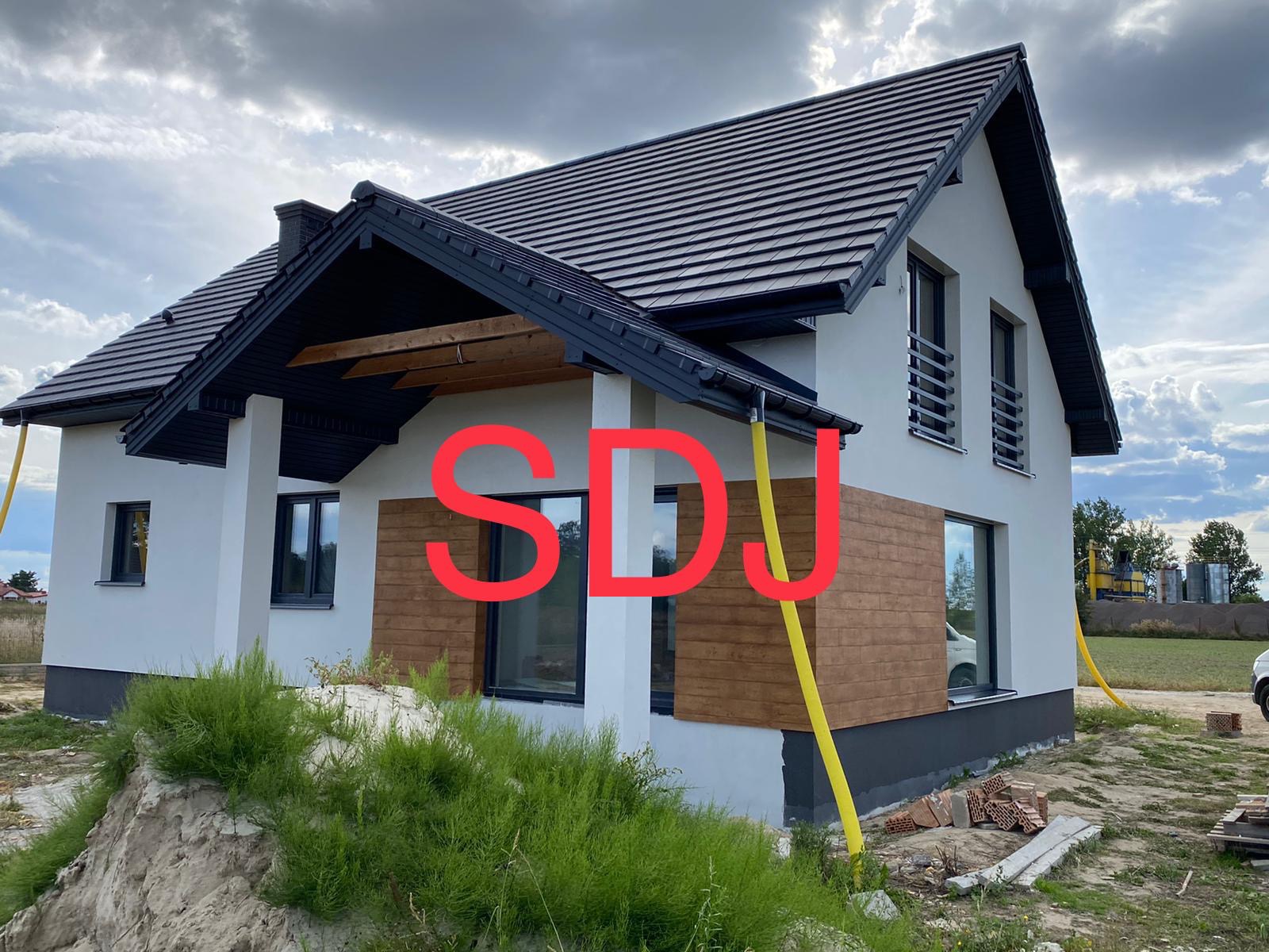 Nowoczesny dom jednorodzinny z białą elewacją, fragmentem drewnianej okładziny, czarnym dachem i orynnowaniem, widoczne elementy rusztowania i teren budowy.