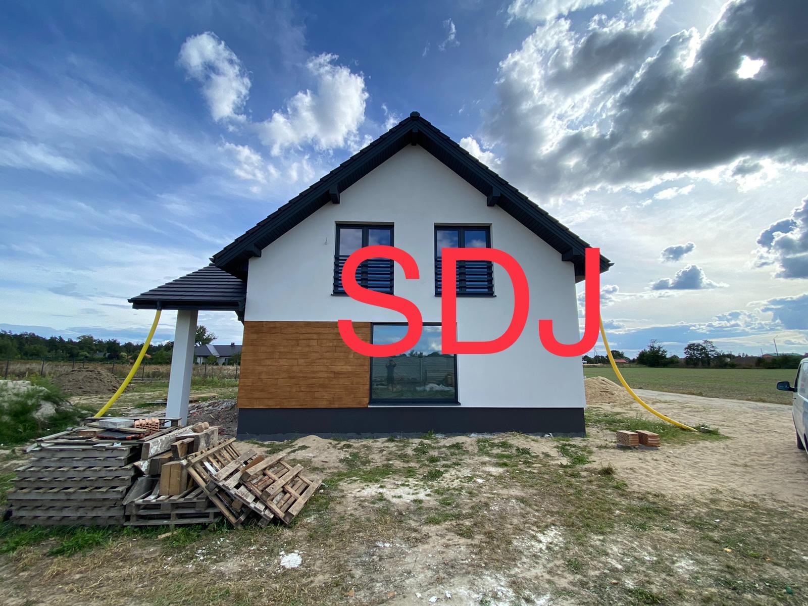 Nowoczesny dom jednorodzinny z fragmentem elewacji wykończonym drewnopodobną okładziną, widoczne palety budowlane i żółty wąż doprowadzający wodę, błękitne niebo z chmurami w tle.