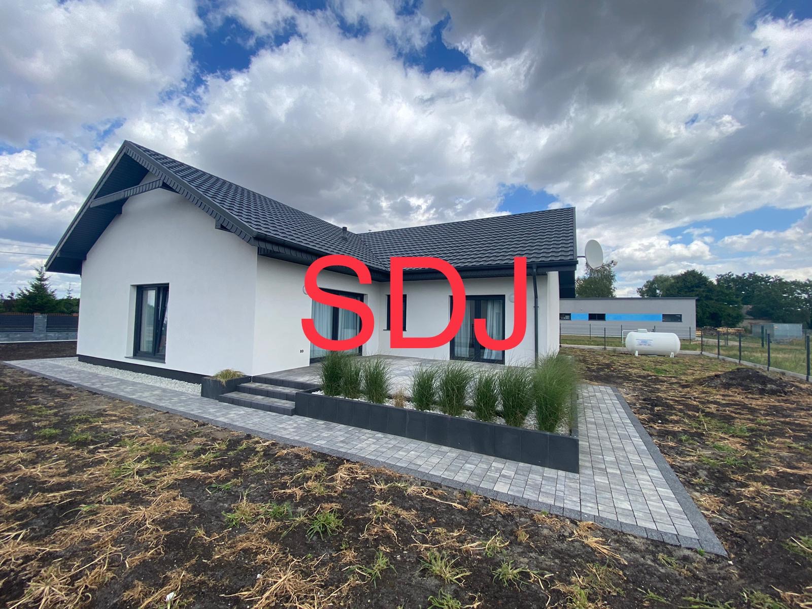 Nowoczesny dom jednorodzinny z białymi ścianami i ciemną dachówką, z utwardzonym terenem wokół, schodami i rabatami z ozdobnymi trawami.