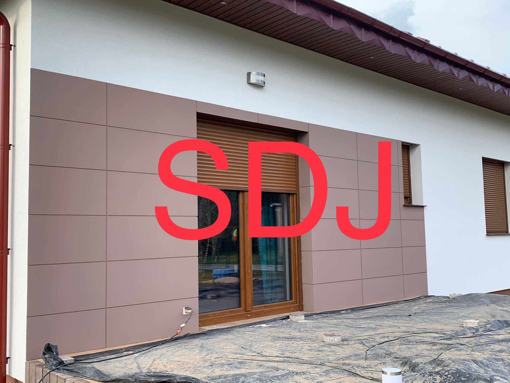 Elewacja budynku z brązowymi panelami, widoczne okno z roletą, teren zabezpieczony folią budowlaną.