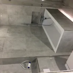 Wykończona łazienka z szarymi płytkami, białą wanną obudowaną płytkami i podświetleniem LED wzdłuż górnej krawędzi, oraz wiszącą toaletą z białą deską. Widoczne są również bateria wannowa i wnęka...