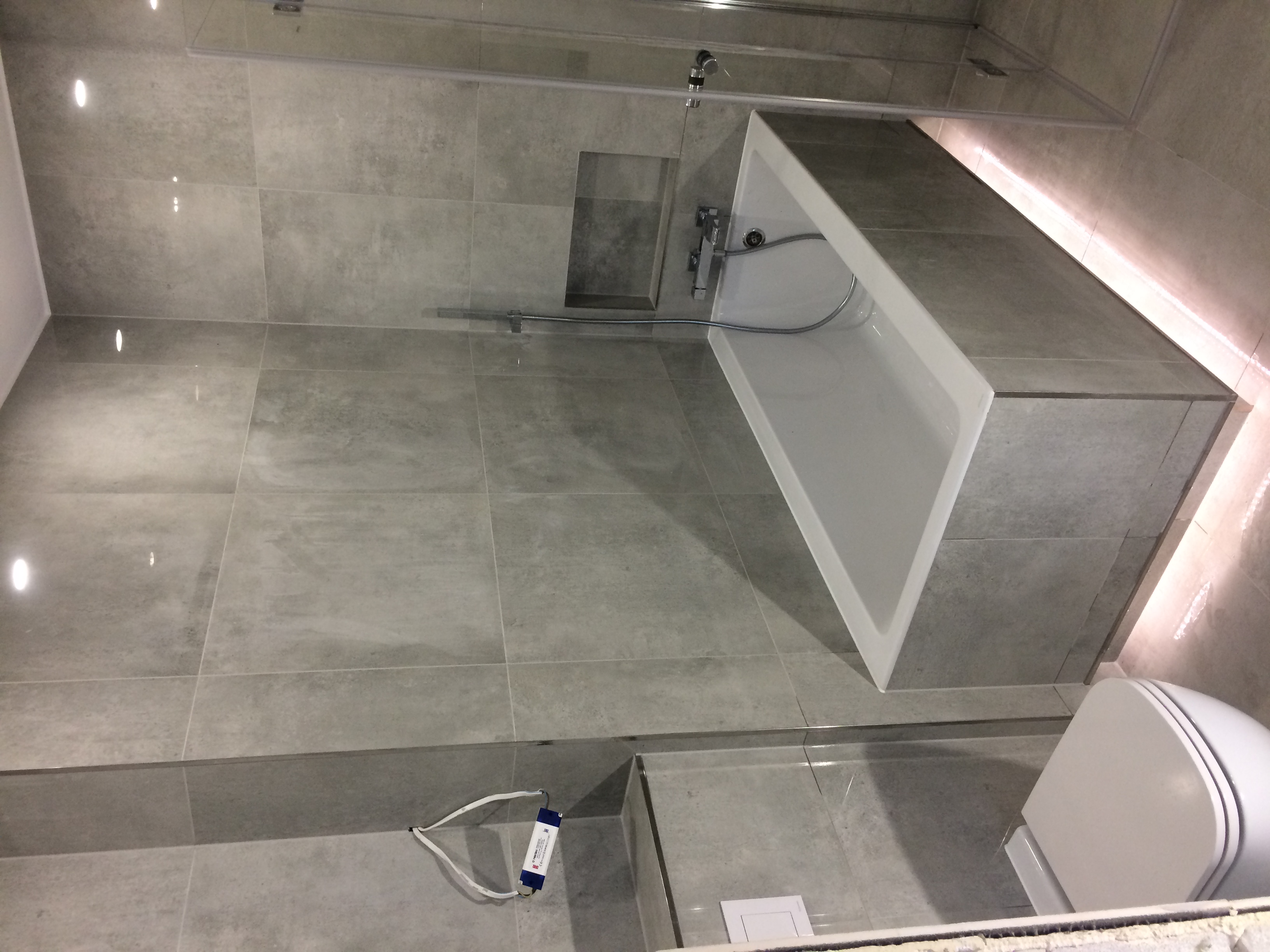 Wykończona łazienka z szarymi płytkami, białą wanną obudowaną płytkami i podświetleniem LED wzdłuż górnej krawędzi, oraz wiszącą toaletą z białą deską. Widoczne są również bateria wannowa i wnęka...