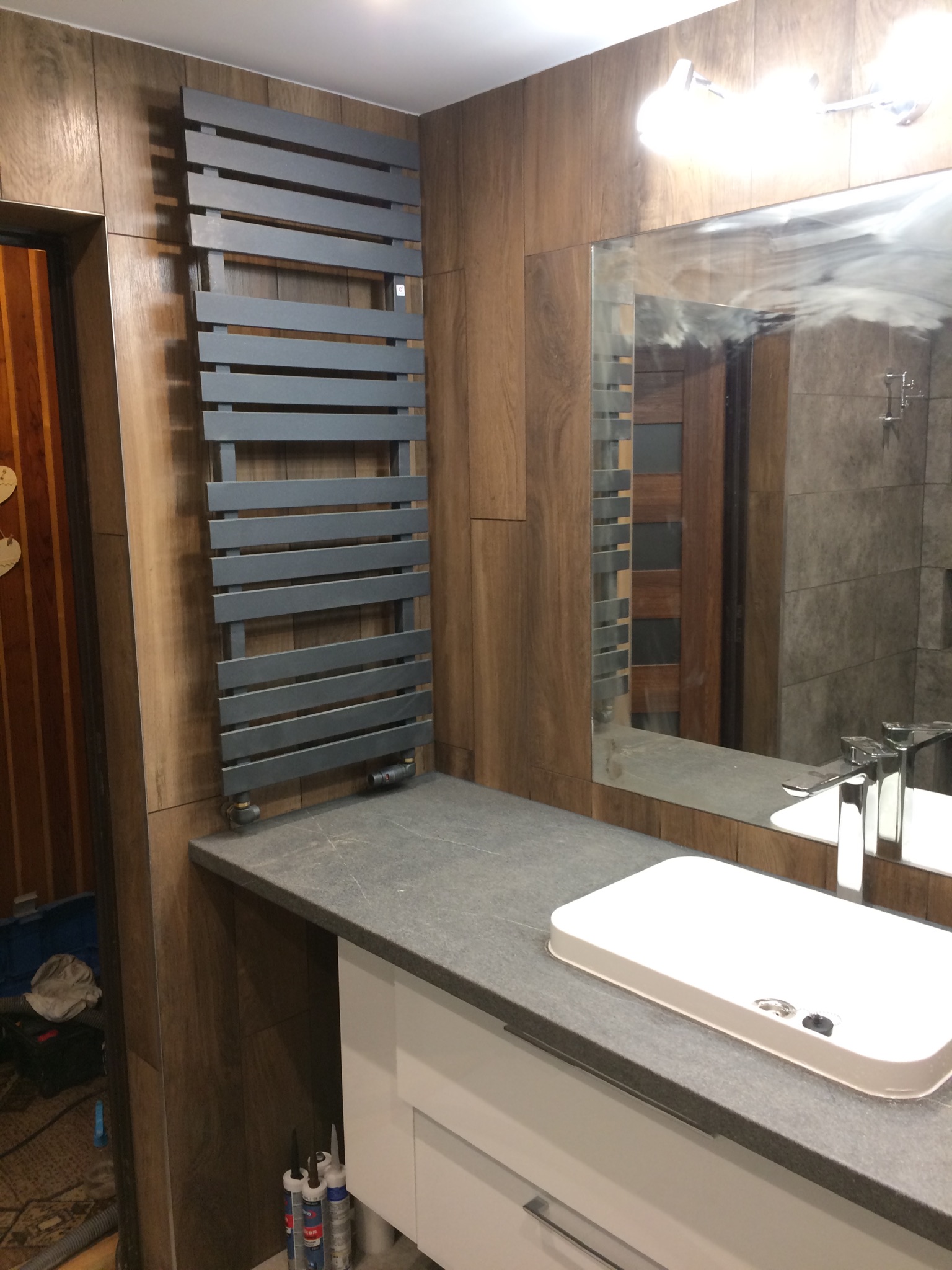 Nowoczesna łazienka z drewnopodobnymi płytkami, szarym grzejnikiem drabinkowym i blatem z umywalką; widoczne tuby silikonu pod blatem.