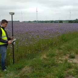 Mężczyzna w żółtej kamizelce odblaskowej obsługuje urządzenie pomiarowe na tle pola kwitnących na fioletowo roślin i turbin wiatrowych w oddali.