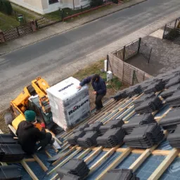 Montaż ciemnoszarej dach&oacute;wki ceramicznej na drewnianej konstrukcji, widoczny podnośnik z paletą dach&oacute;wek marki Roben, praca wykonywana przez dw&oacute;ch dekarzy na tle zabudowań mieszkalnych.