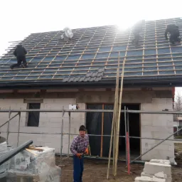 Budowa domu jednorodzinnego: ekipa montuje łatowanie pod pokrycie dachu, część dachówek leży na folii, na pierwszym planie pracownik w czapce i pomarańczowych rękawicach.