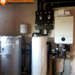 Nowoczesna kotłownia z dwoma zbiornikami na wodę (jeden w folii ochronnej), białym kotłem Bosch, czarną armaturą i rurami, na tle surowej ściany z widocznymi śladami montażu.