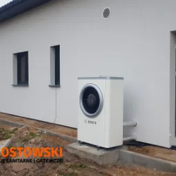 Biała pompa ciepła Bosch zamontowana przy ścianie domu z szarymi oknami i drzwiami, widoczne rury przyłączeniowe, teren wokół częściowo zagospodarowany.