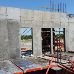 Wznoszenie betonowej ściany szalunkowej z otworami okiennymi, widoczne zbrojenie i elementy konstrukcyjne, w tle pole kukurydzy.