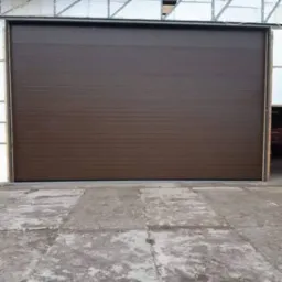 Brązowa brama garażowa segmentowa z poziomymi przetłoczeniami, widoczna na tle szarej betonowej posadzki i białych ścian budynku.