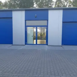 Nowoczesny budynek przemysłowy z niebieskimi bramami rolowanymi i przeszklonym wejściem, wykończony panelami w kolorze białym i niebieskim, z kostką brukową na pierwszym planie.