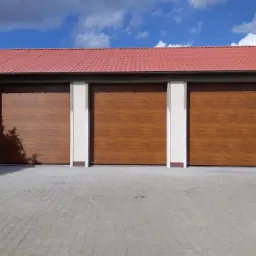 Trzy brązowe bramy garażowe segmentowe z poziomym wzorem drewna, zamontowane w jasnym budynku z czerwoną dach&oacute;wką, widoczne z zewnątrz na brukowanym podjeździe.