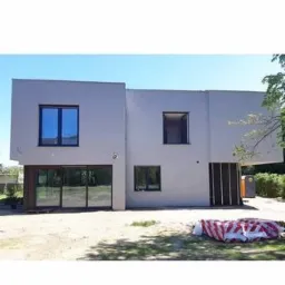 Nowoczesny, dwukondygnacyjny dom jednorodzinny z płaskim dachem, wykończony jasnoszarym tynkiem, z dużymi przesuwnymi drzwiami tarasowymi i prostokątnymi oknami o ciemnych ramach. Na pierwszym...