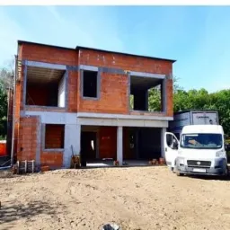 Dwukondygnacyjny dom w trakcie budowy z czerwonej cegły, widoczne surowe otwory okienne i drzwiowe, na pierwszym planie piaszczysty teren budowy, z boku zaparkowany biały bus z otwartymi drzwiami.