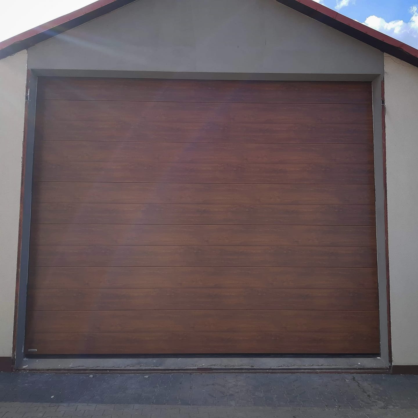 Zamknięta, segmentowa brama garażowa w kolorze ciemnego drewna, zamontowana w szarej bryle budynku z widocznym fragmentem dachu i kostką brukową przed bramą.