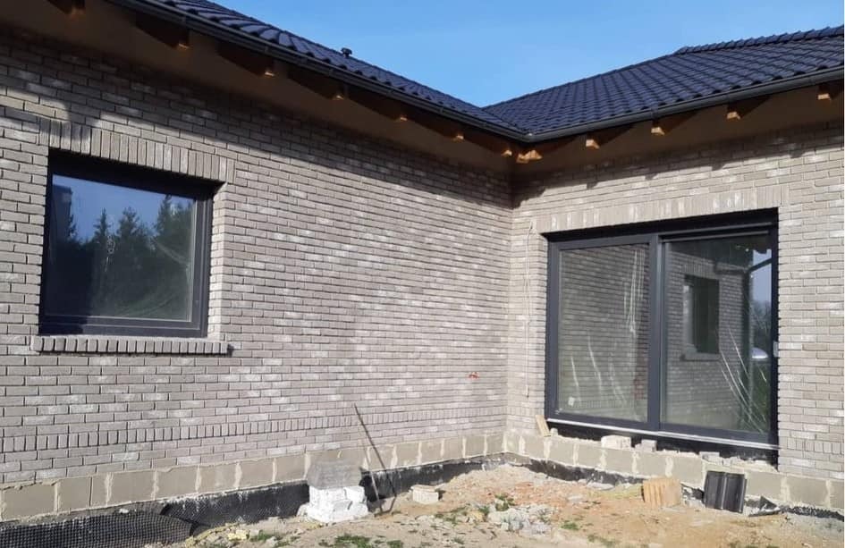 Szary budynek z cegły klinkierowej w trakcie budowy, z zamontowanymi oknami z ciemnymi ramami i widocznymi elementami izolacji fundamentów oraz fragmentem dachu z ciemnej dachówki.