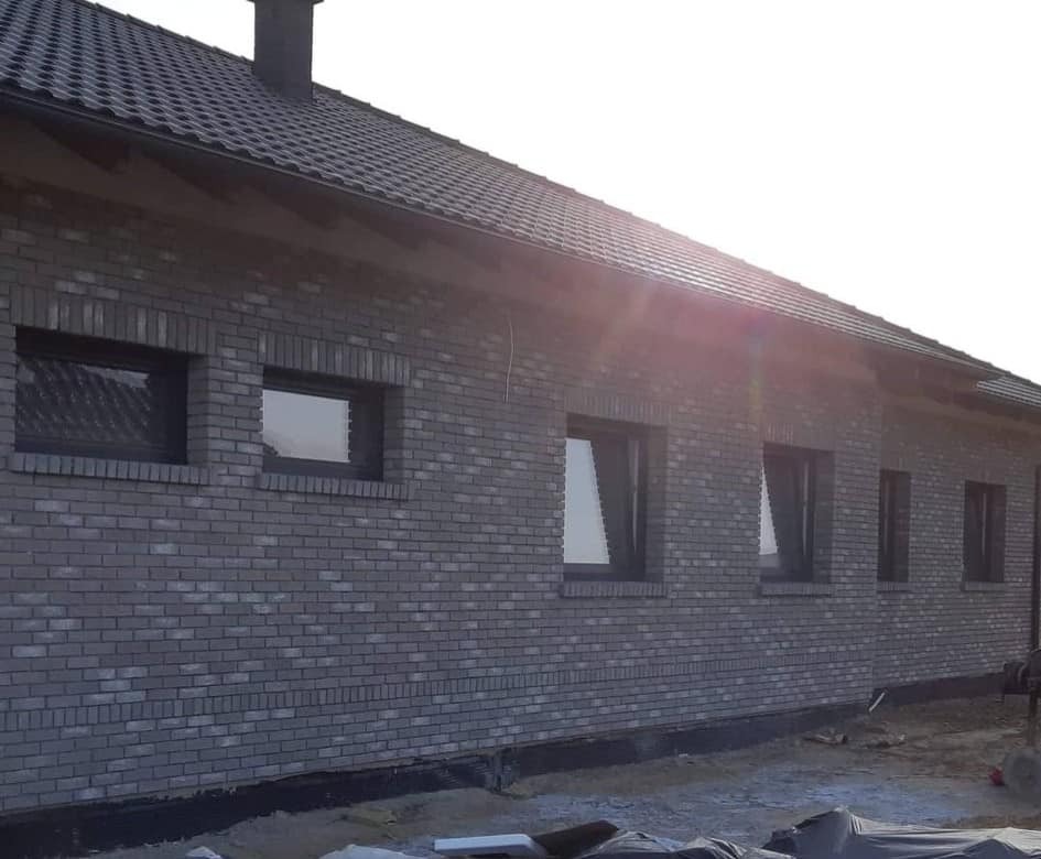 Szary dom z cegły klinkierowej z zamontowanymi oknami z czarnymi ramami, widok z zewnątrz z lekkim prześwietleniem słonecznym.