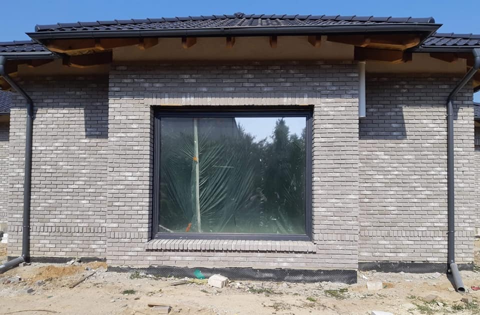 Szara ściana z cegły klinkierowej z zamontowanym oknem, zabezpieczonym folią ochronną, widoczne rynny i fragment dachu z ciemnej dachówki.