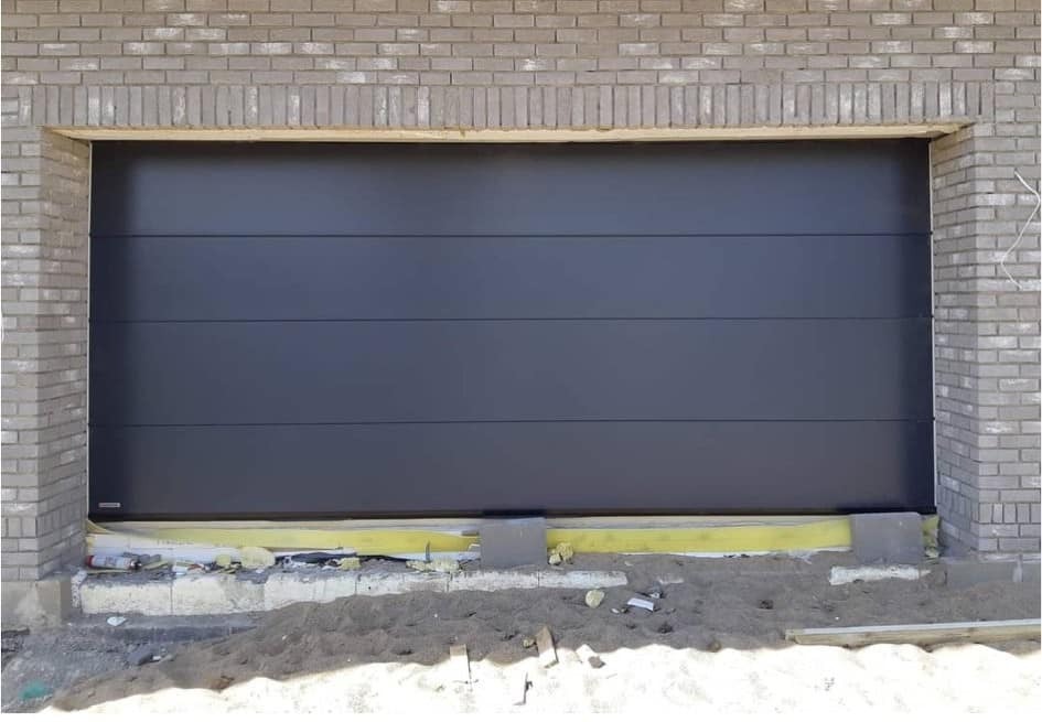 Nowoczesna, segmentowa brama garażowa w kolorze grafitowym, zamontowana w surowym murze z jasnej cegły, widoczne ślady prac budowlanych i piasek na posadzce.
