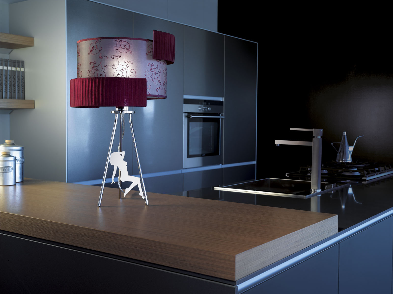 Nowoczesna lampa stołowa z abażurem w odcieniach czerwieni i wzorami, na metalowym statywie z wyciętą sylwetką tancerki, na blacie kuchennym w minimalistycznej kuchni z ciemnymi szafkami.