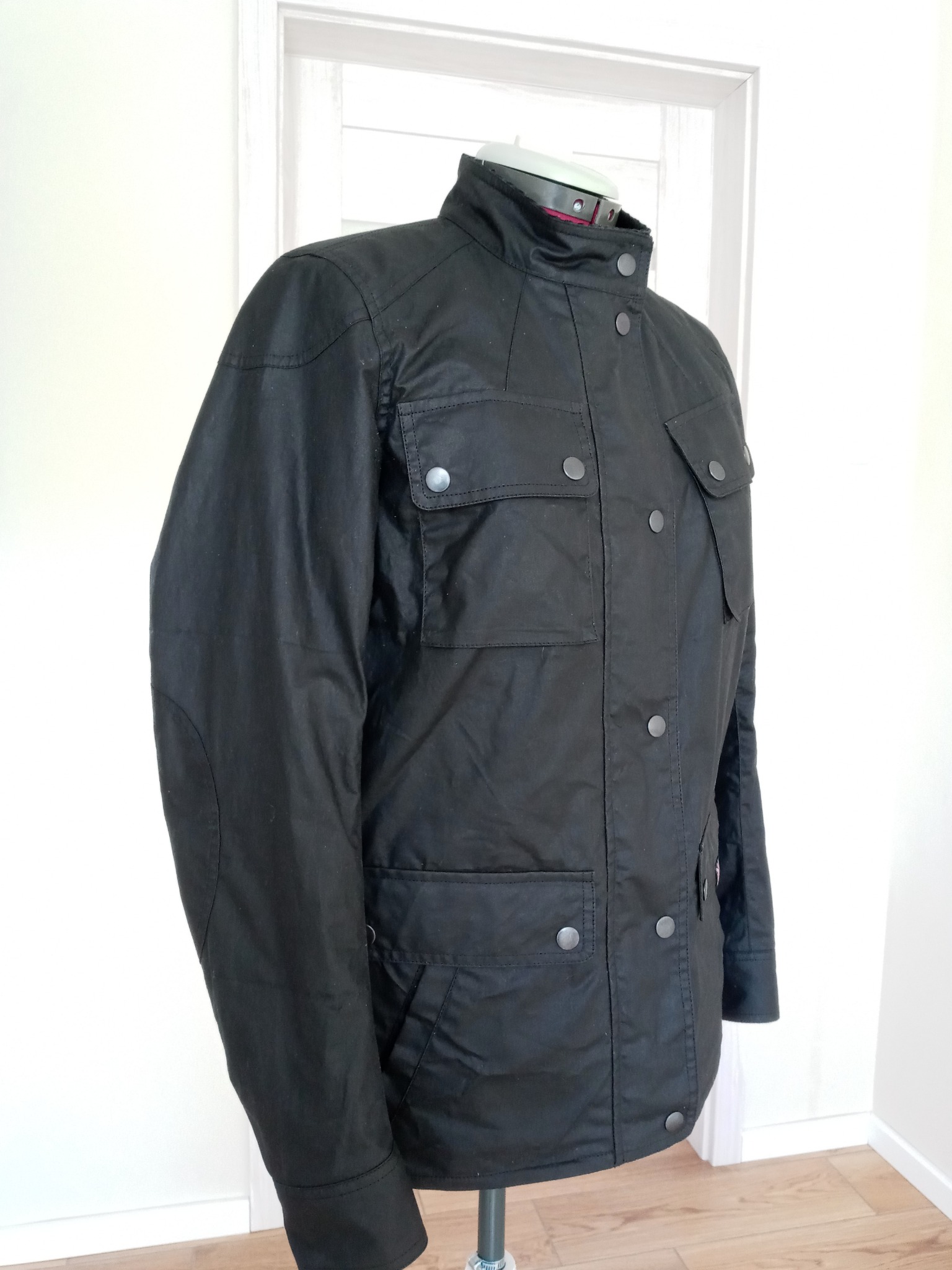 Czarna, woskowana kurtka na manekinie, widoczne kieszenie z klapami i metalowe zatrzaski, jasne tło z drzwiami.