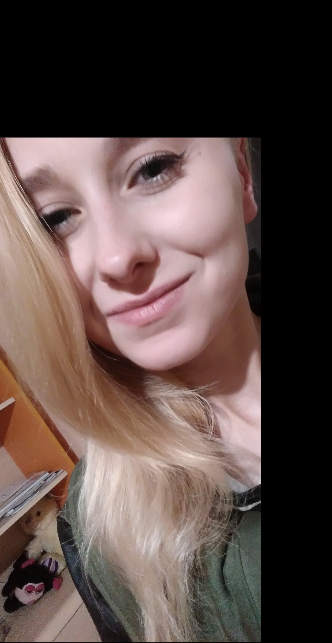 Portret uśmiechniętej, młodej blondynki z delikatnym makijażem, w tle fragment półki z zabawkami.