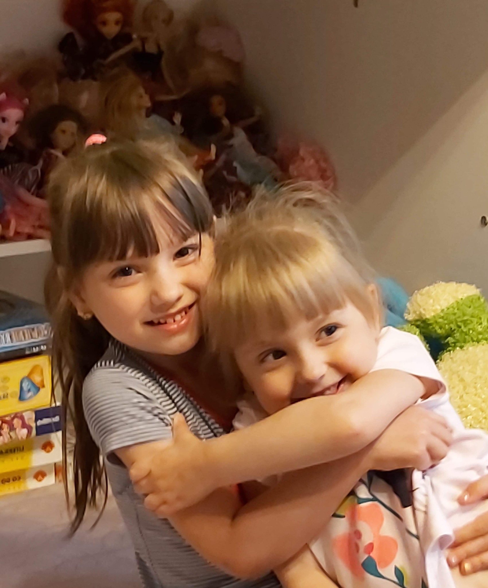 Dwie uśmiechnięte dziewczynki obejmujące się na tle półki z lalkami i książkami.