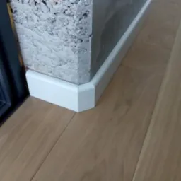 Fragment drewnianej podłogi z przypodłogową listwą w kolorze białym, narożnik ściany z surowego betonu oraz fragment czarnej ramy drzwiowej.