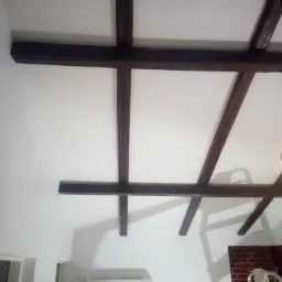 Biały sufit z dekoracyjnymi, ciemnymi belkami drewnianymi, klimatyzator na ścianie, fragment ściany z cegły w tle.