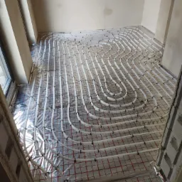 Białe rury ogrzewania podłogowego ułożone na folii izolacyjnej z czerwoną siatką, przymocowane czarnymi klipsami w surowym wnętrzu budynku w trakcie budowy.