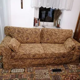 Dwuosobowa sofa z brązową tapicerką w roślinny wzór, na tle okna z firanką i drewnianej witryny z porcelaną, na wzorzystym dywanie.